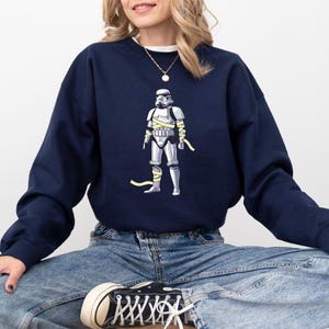 May include: Navy blue sweatshirt featuring a graphic of a Stormtrooper wrapped in yellow bandages. The Stormtrooper is in a standing position. The sweatshirt is a solid navy blue color.