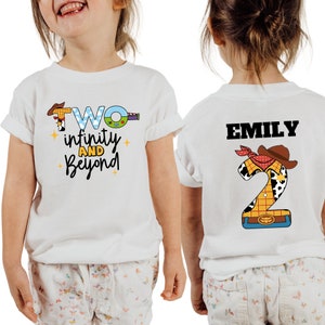 May include: Two white t-shirts with a Toy Story theme. The front of one shirt says "Two infinity and beyond" with a cartoon image of Woody. The back of the other shirt has a large number "2" with a cartoon image of Woody and the name "EMILY" above it.