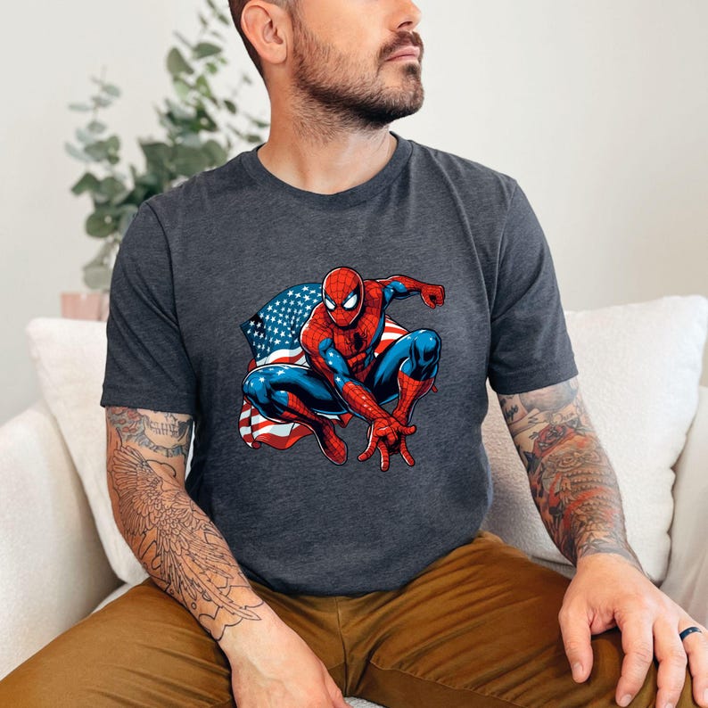 May include: Dark gray t-shirt featuring a graphic of Spider-Man in a crouching pose with the American flag in the background. The superhero is depicted in his classic red and blue suit.