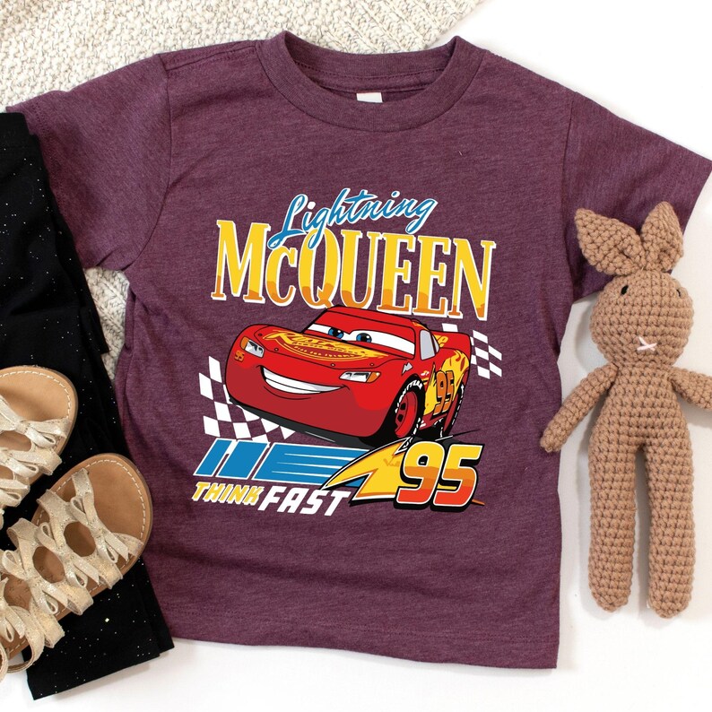 Puede incluir: Una camiseta granate con un dise&ntilde;o de Rayo McQueen de la pel&iacute;cula Cars. El gr&aacute;fico incluye el texto "Lightning McQueen", el n&uacute;mero "95" y la frase "Think Fast". Tambi&eacute;n se ven unos pantalones negros, sandalias doradas y un conejito de peluche marr&oacute;n.