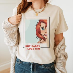 May include: A beige t-shirt featuring a cartoon image of a red-haired woman with blue eyes and a red mouth. The text "BUT DADDY I LOVE HIM" is printed below the image.