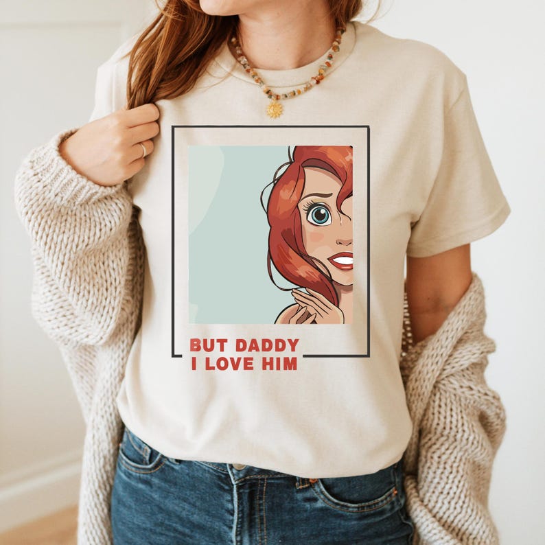 May include: A beige t-shirt featuring a cartoon image of a red-haired woman with blue eyes and a red mouth. The text "BUT DADDY I LOVE HIM" is printed below the image.