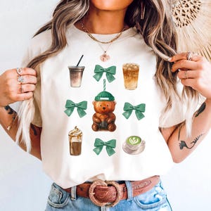 May include: A cream-colored t-shirt featuring a collection of watercolor illustrations. The design includes various coffee drinks, green bows, and a brown teddy bear wearing a green beanie. The shirt is a casual, everyday wear item.