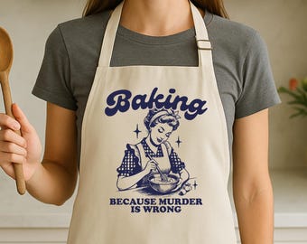 Pode incluir: Avental creme com a palavra "Baking" em escrita azul marinho. Abaixo, uma ilustração vintage de uma mulher batendo em uma tigela, com o texto "BECAUSE MURDER IS WRONG". O avental tem um babador e uma alça de pescoço.
