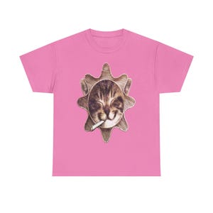 May include: Pink t-shirt with a graphic of a cat's face smoking a cigarette, set within a sun-like design. The cat's face is brown and tan, with closed eyes. The shirt is made of a soft material.