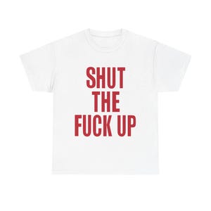 Puede incluir: Camiseta blanca con la frase "SHUT THE FUCK UP" impresa en letras rojas en negrita. El texto está centrado en la parte delantera de la camiseta. La camiseta tiene mangas cortas y cuello redondo.