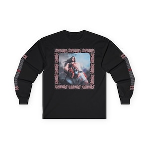 May include: Black long-sleeve shirt featuring a central graphic of a muscular figure with long hair, holding a heart. The graphic is framed by ornate text. Additional text adorns the sleeves. The shirt is made of a soft material.