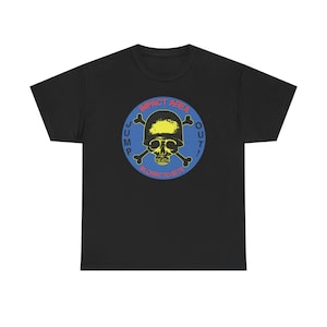 Puede incluir: Camiseta negra con un diseño circular. El diseño incluye una calavera con huesos cruzados, un casco amarillo y las palabras "IMPACT AREA" y "BLOWN TO BITS". El diseño es en azul, amarillo y rojo.