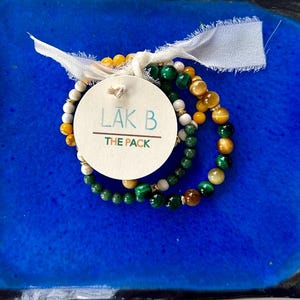 May include: Three beaded bracelets in shades of green, yellow, and white are displayed on a square, cobalt blue ceramic plate. A round tag with the text "LAK B THE PACK" is attached with a white ribbon. The plate has a dark blue border.