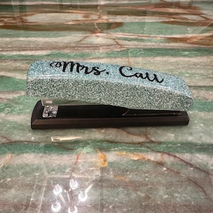 Może przedstawiać: Turkusowa zszywaczka z brokatem z czarną podstawą. Górna część zszywaczki jest ozdobiona napisem "Mrs. Catt" czarnymi literami.