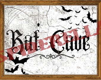 May include: A framed print with a Halloween theme. The artwork features the words "Bat Cave" in a gothic font, along with illustrations of bats, a spider, and a spiderweb. The background has a distressed, textured appearance.