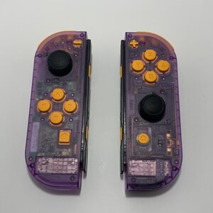 May include: Two transparent purple and orange game controllers. Each controller has a black joystick and orange buttons. The controllers are designed for a gaming console. The buttons are labeled with letters and symbols.