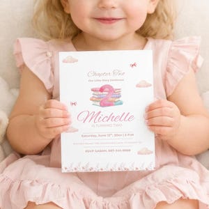 May include: A young child in a pink dress holds a custom invitation. The invitation features the name "Michelle" in cursive, with illustrations of books and clouds. The child is seated in a cream-coloured chair, surrounded by pink bows and a stuffed bunny.