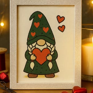 Può includere: Opera d'arte incorniciata con uno gnomo che tiene un cuore rosso. Lo gnomo indossa un cappello verde con cuori rossi e un cappotto verde. Due cuori rossi aggiuntivi sono accanto allo gnomo. La cornice è argentata e lo sfondo è beige chiaro.