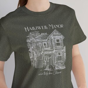 Pode incluir: Uma camiseta verde escura com um gráfico branco de uma casa e o texto "Halliwell Manor" e "we've truly been blessed".