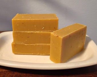May include: Four rectangular bars of yellow soap are stacked on a white, oval plate. The soap has a slightly rough texture and a warm, golden colour. One bar stands separately, next to the stack. The background is a light blue.