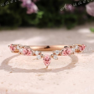 Puede incluir: Un anillo de oro rosa con un diseño curvo, con piedras preciosas rosas y pequeños diamantes blancos. El anillo tiene una piedra rosa central en forma de pera, sobre un fondo neutro.