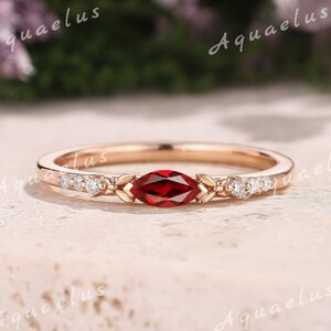 May include: Rose gold ring with a marquise-cut, deep red gemstone centre, leaf-like designs, and small accent stones. The delicate, polished band is set against a neutral background. The ring is a beautiful piece of jewellery.