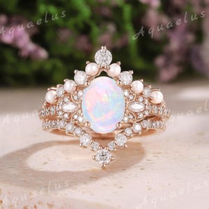 May include: A rose gold engagement ring set with a large oval opal surrounded by smaller diamonds and pearls. The ring has a delicate, vintage-inspired design.