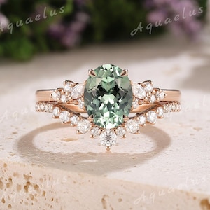 Könnte beinhalten: Ein Verlobungsring aus Roségold mit einem grünen ovalen Edelstein, der von kleineren Diamanten umgeben ist. Der Ring hat ein zartes, florales Design.