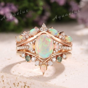 Könnte beinhalten: Ein dreiteiliger Verlobungsring aus Roségold mit einem großen ovalen Opal als Mittelstück und kleineren Opal-Akzenten. Der Ring ist mit funkelnden Diamanten und grünen Edelsteinen verziert.