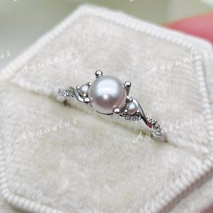Puede incluir: Un anillo de tono plateado con una perla blanca redonda central flanqueada por dos perlas más pequeñas a cada lado. La banda tiene un diseño trenzado con pequeñas piedras transparentes incrustadas en el metal, dentro de una caja hexagonal crema.