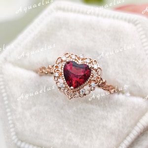May include: A rose gold ring featuring a central heart-shaped red gemstone surrounded by a decorative filigree frame accented with small clear stones. The band has a twisted metal texture. The word Aquaelus is repeated across the image.