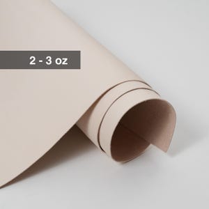 May include: A rolled sheet of light beige leather. The text "2 - 3 oz" is displayed on a gray rectangle in the upper left corner. The leather is smooth and appears to be new.