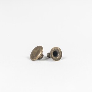 May include: Two antique brass rivets, each with a rounded cap and a hollow post. The rivets are a warm, metallic color, and are set against a plain white background. These rivets are likely used for leathercraft or garment construction.