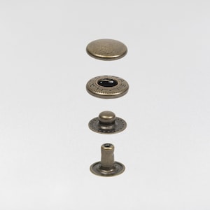 May include: An assortment of antique brass-colored snap fasteners, disassembled and arranged vertically. The components include a domed cap, a ring with embossed text, a stud, and a post. The fasteners are designed for securing fabric or other materials.