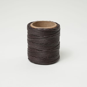 May include: A spool of dark brown waxed thread, tightly wound around a cardboard core. The thread appears smooth and durable, suitable for various crafting projects. The spool is set against a plain white background.