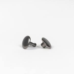 May include: Two dark gray oval-shaped rivets with short stems, set against a white background. The rivets have a smooth, metallic finish and are slightly angled, showcasing their rounded form. These fasteners are likely used in leathercraft or garment construction.