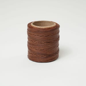 May include: A spool of brown leather cord, tightly wound around a cardboard core. The cord appears to be made of thin, flat strips, creating a textured surface. The color is a warm, medium brown.