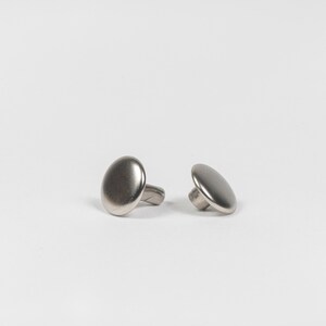 May include: Two silver-toned oval cufflinks on a white background. One cufflink is upright, while the other is lying on its side, showcasing the post. The cufflinks have a brushed metal finish.