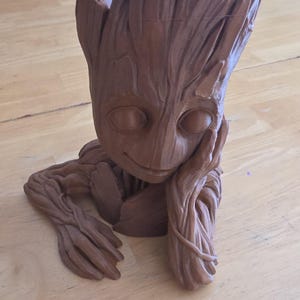 May include: A brown, stylized Groot planter, featuring a smiling face and arms resting on a surface. The planter has a textured, wood-like appearance, with a hollowed-out top for plants. A charming and unique decorative item.