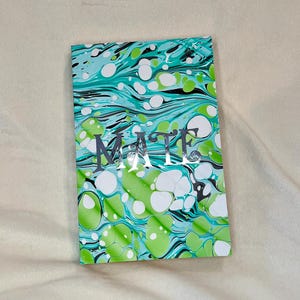 Puede incluir: Un cuaderno con un diseño veteado en tonos verdes, blancos y turquesas. La palabra "MATTE" está impresa en plata en la portada. El cuaderno rectangular descansa sobre una superficie beige.