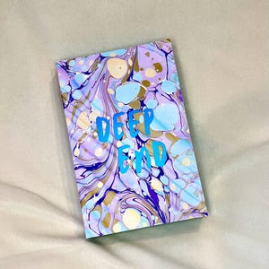 May include: A rectangular notebook with a marbled design in shades of purple, blue, and gold. The words "DEEP END" are written in bright blue, centered on the cover. The notebook is lying on a light gray surface.