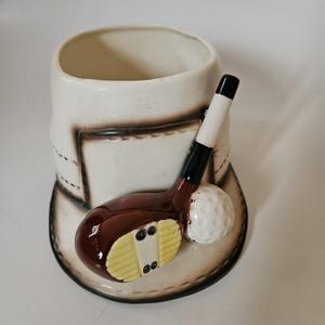 May include: A white ceramic golf-themed mug with a brown golf club and a white golf ball. The mug has a brown rim and a brown stitched design around the top.