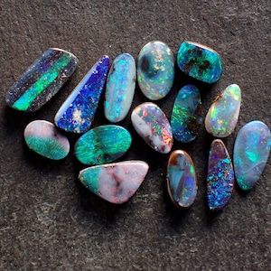 May include: A collection of polished opal gemstones in various shapes and sizes. The opals display a range of colors, including blues, greens, and purples, with iridescent patterns. The gemstones are arranged on a dark, textured surface.