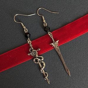 Puede incluir: Un par de pendientes de plata con detalles de cristal negro. Un pendiente presenta un diseño de espada, mientras que el otro presenta una serpiente envuelta alrededor de una espada.