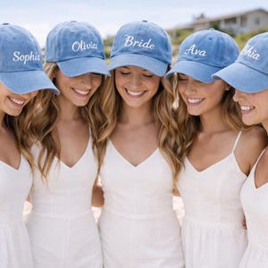 May include: Five people wearing matching light blue baseball caps with white embroidered names and the word "Bride". They are smiling and wearing white dresses. The photo is taken outdoors on a sunny day.