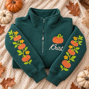 Pode incluir: Uma camisola verde-escura com fecho de correr curto, apresentando abóboras cor de laranja e flores amarelas bordadas nas mangas. No peito esquerdo, uma abóbora cor de laranja bordada está sobre o texto cursivo branco que diz Krist.
