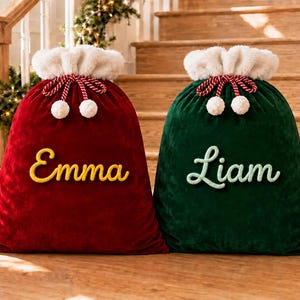Pode incluir: Dois grandes sacos de presente de veludo com punhos de pele sintética branca e fechos de cordão com pompons. O saco vermelho tem o nome Emma bordado em amarelo e o saco verde tem o nome Liam bordado em letras cursivas verde-claras.