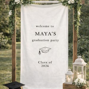 Puede incluir: Un letrero de fiesta de graduación con el texto "welcome to MAYA'S graduation party" y "Class of 2026". El letrero está decorado con flores y vegetación. Un birrete y un pergamino están en la base.