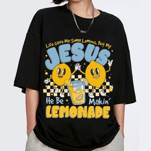 Può includere: T-shirt nera con un design grafico raffigurante due limoni dei cartoni animati, un bicchiere di limonata e il testo "Life Gave Me Some Lemons, But My JESUS He Be Makin' LEMONADE" in blu e giallo.