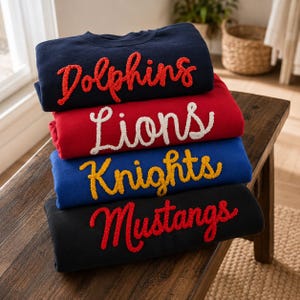 May include: A stack of four crewneck sweatshirts in navy, red, blue, and black. Each sweatshirt features textured, raised lettering in a cursive font. The navy sweatshirt says Dolphins in red, red says Lions in white, blue says Knights in yellow, and black says Mustangs in red.