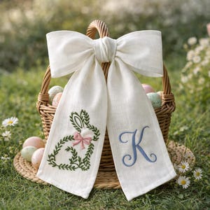 May include: A white linen Easter bow with long streamers. One streamer features a green embroidered bunny with a pink bow, the other has a blue embroidered "K". A wicker basket filled with Easter eggs is in the background.