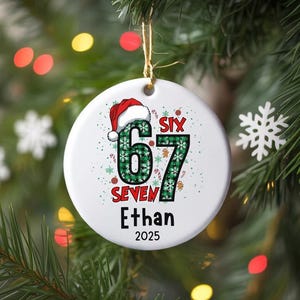 May include: A white Christmas ornament with the text "SIX 67 SEVEN Ethan 2025". The numbers are in a green plaid pattern with a Santa hat on the number 6. The ornament is hanging on a Christmas tree.