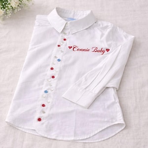 Może przedstawiać: Biała koszula zapinana na guziki z klasycznym kołnierzykiem i długimi rękawami. Koszula posiada rząd kolorowych guzików i czerwony haft z napisem "Connie Baby" z symbolami serca. Stylowy i spersonalizowany element garderoby.
