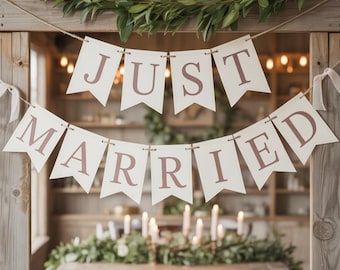 May include: A cream-coloured banner with the words "JUST MARRIED" in mauve lettering. The banner is suspended from a wooden frame, with a table set with candles and greenery in the background, creating a romantic scene.